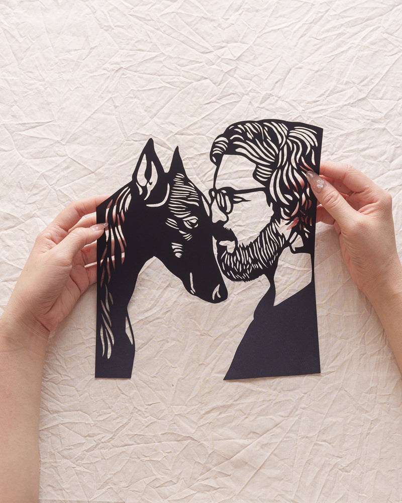 A hand holds a finely crafted black paper-cut artwork featuring a Doberman Pinscher and its owner with their heads touching gently.