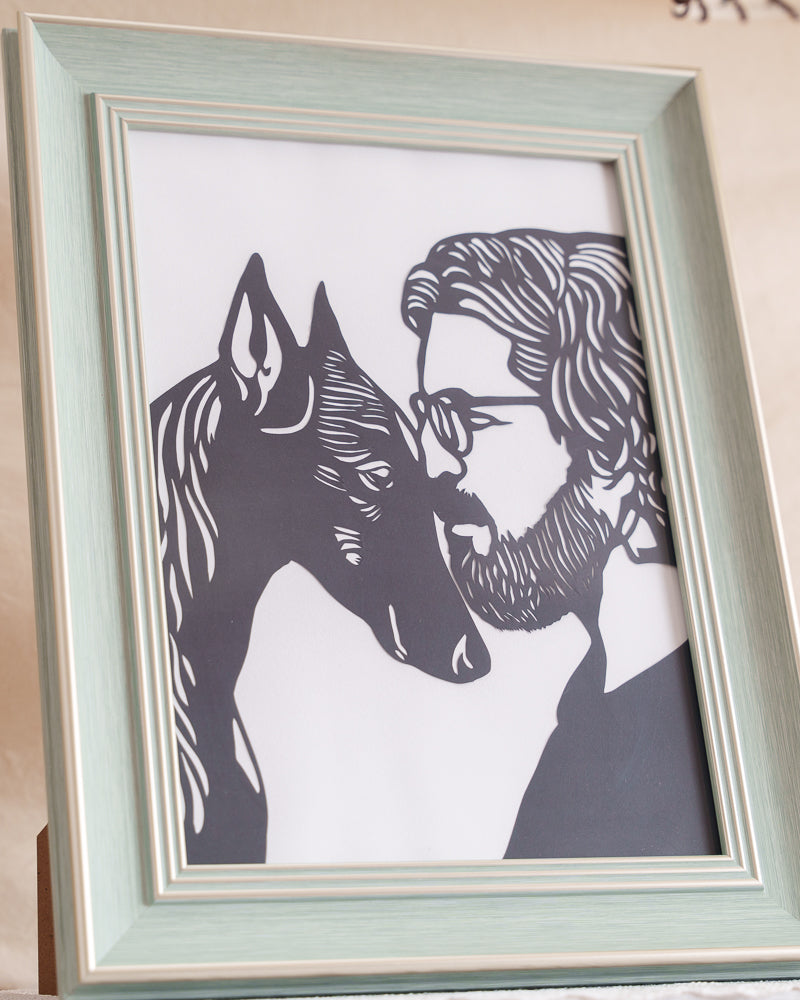 Close-up detail of a black paper-cut artwork, showcasing the precise cut lines and elegant silhouettes of a Doberman Pinscher and its owner touching heads.
