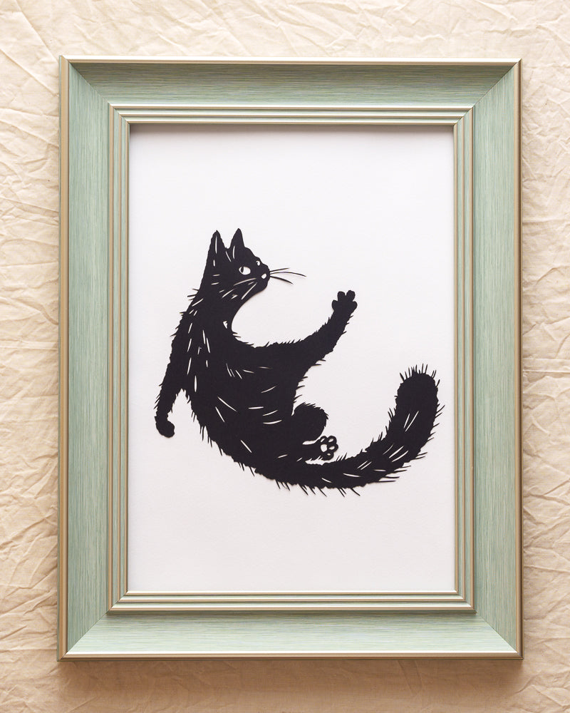 Hand-cut silhouette of kitten stretching with front paws grounded, hind legs lifted and curly tail raised on light gray background