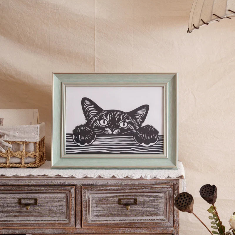 A papercut artwork displayed at home, depicting a cat lying on a table, sneakily peeking over the edge — capturing a playful and curious moment.