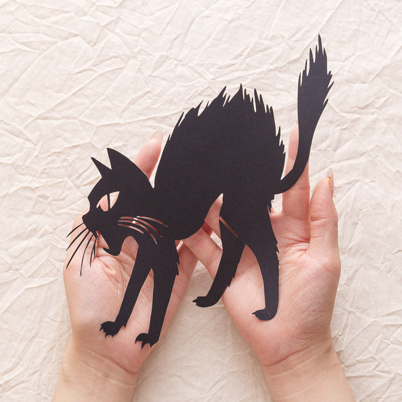 A hand holding a handcrafted black cat paper cut, capturing the startled expression and pose of the cat.