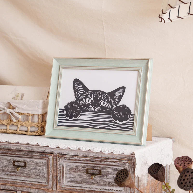 A papercut artwork displayed at home, depicting a cat lying on a table, sneakily peeking over the edge — capturing a playful and curious moment.